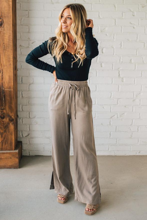 Woman wearing a black top and beige high-waisted pants against a white brick wall.