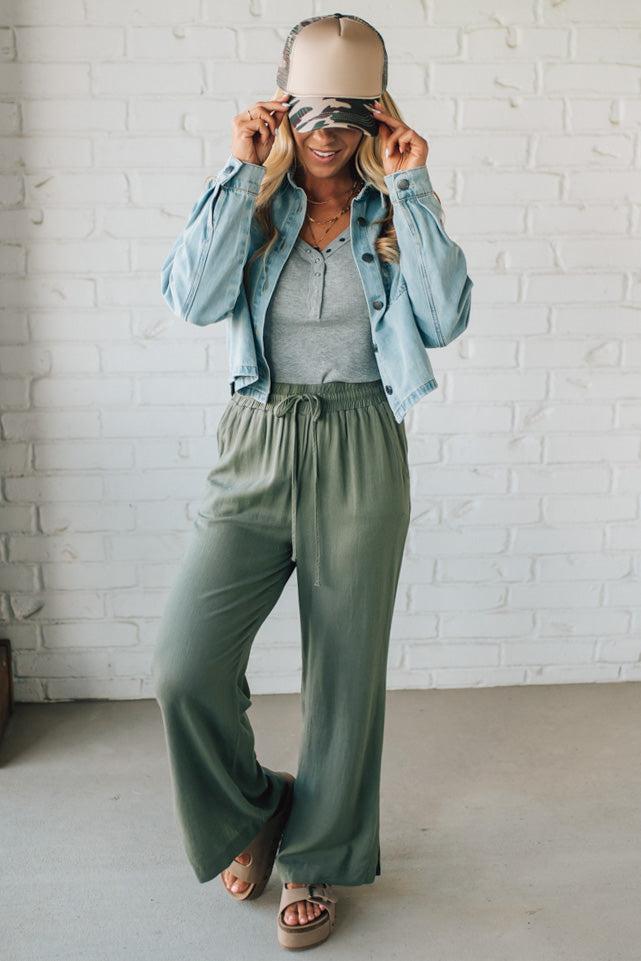 Woman wearing a denim jacket, gray top, and green pants against a white brick wall.