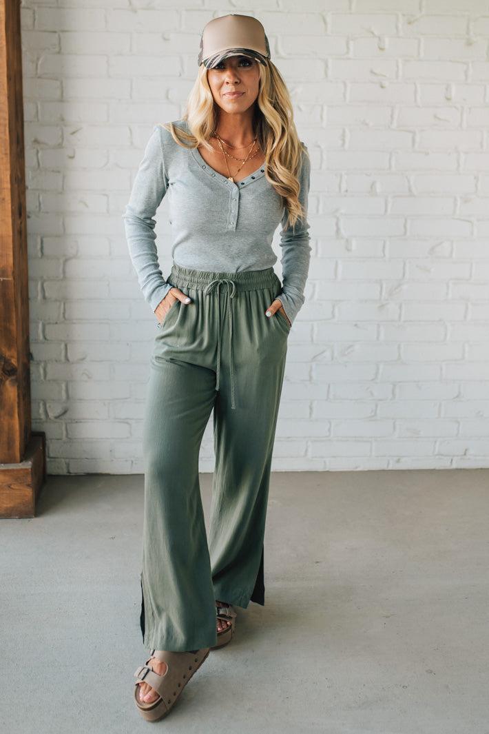 Woman wearing a light gray long-sleeve top and green wide-leg pants against a white brick wall.