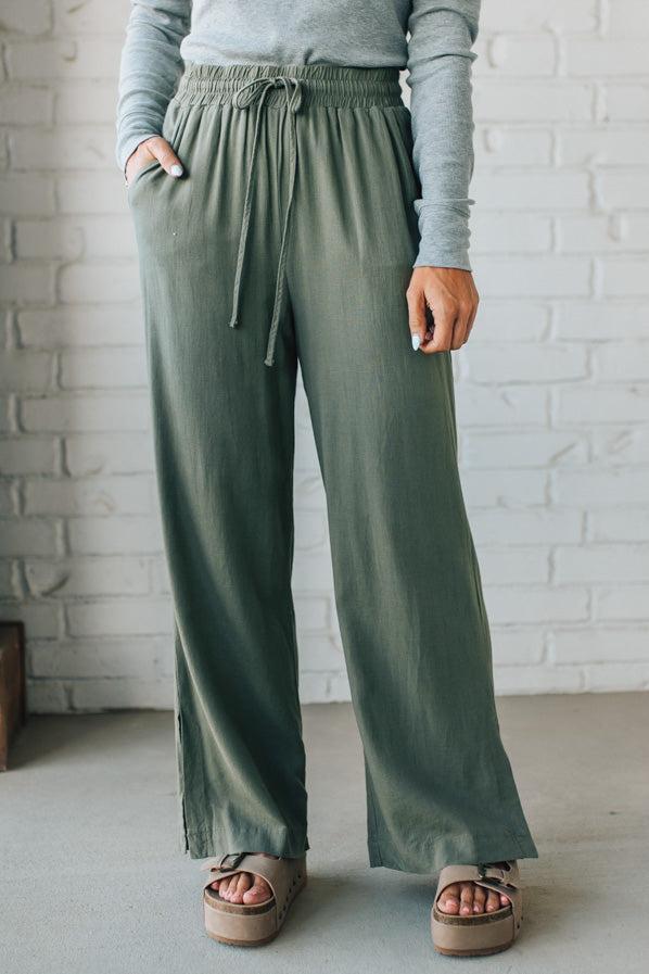 Person wearing green wide-leg pants and a gray long-sleeve top against a white brick wall.