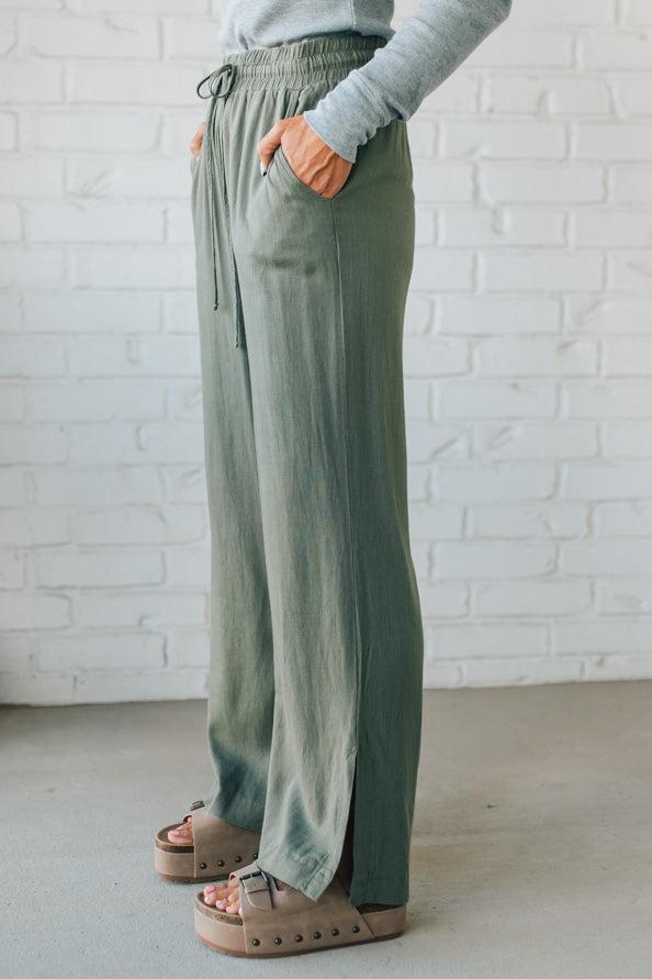 Person wearing green wide-leg pants and a gray top against a white brick wall.