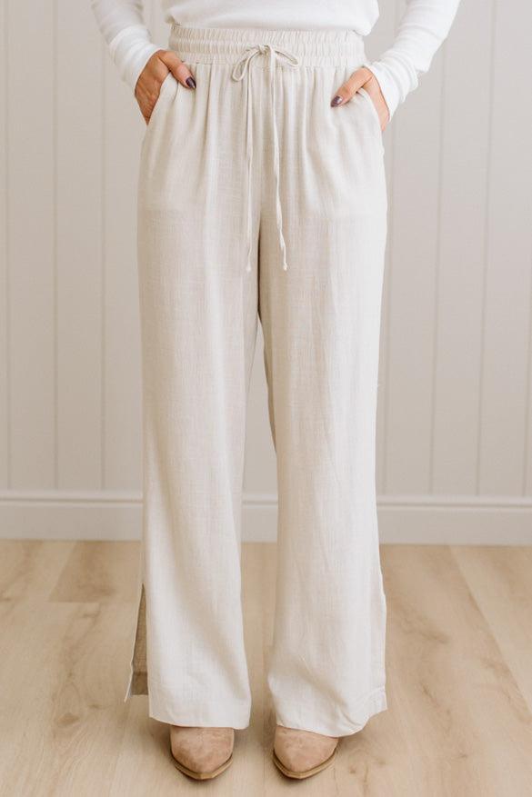 Person wearing beige wide-leg pants and a white top against a light wooden floor and wall background.