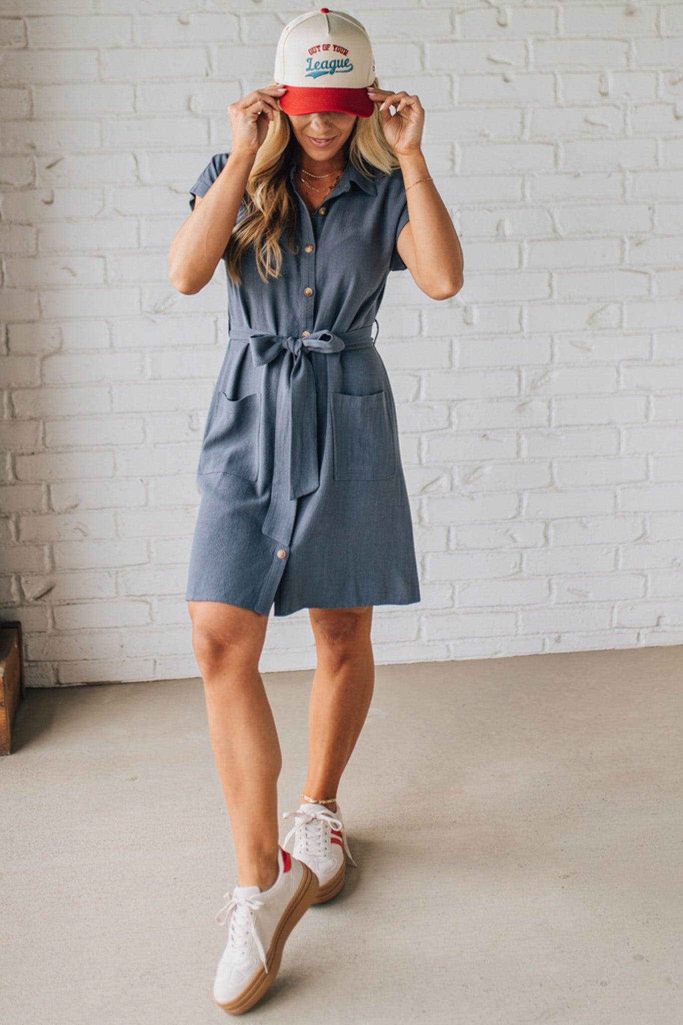 BLONDE WOMAN WARING A KNEE LENGTH SHORT DRESS WITH BUTTON UP AND COLLARED FRONT. HAS SHORT SLEEVES, FRONT PATCH POCKETS AND TIES AT THE WAIST, ALL IN SLATE BLUE WITH A TEXTURED LINEN BLEND.