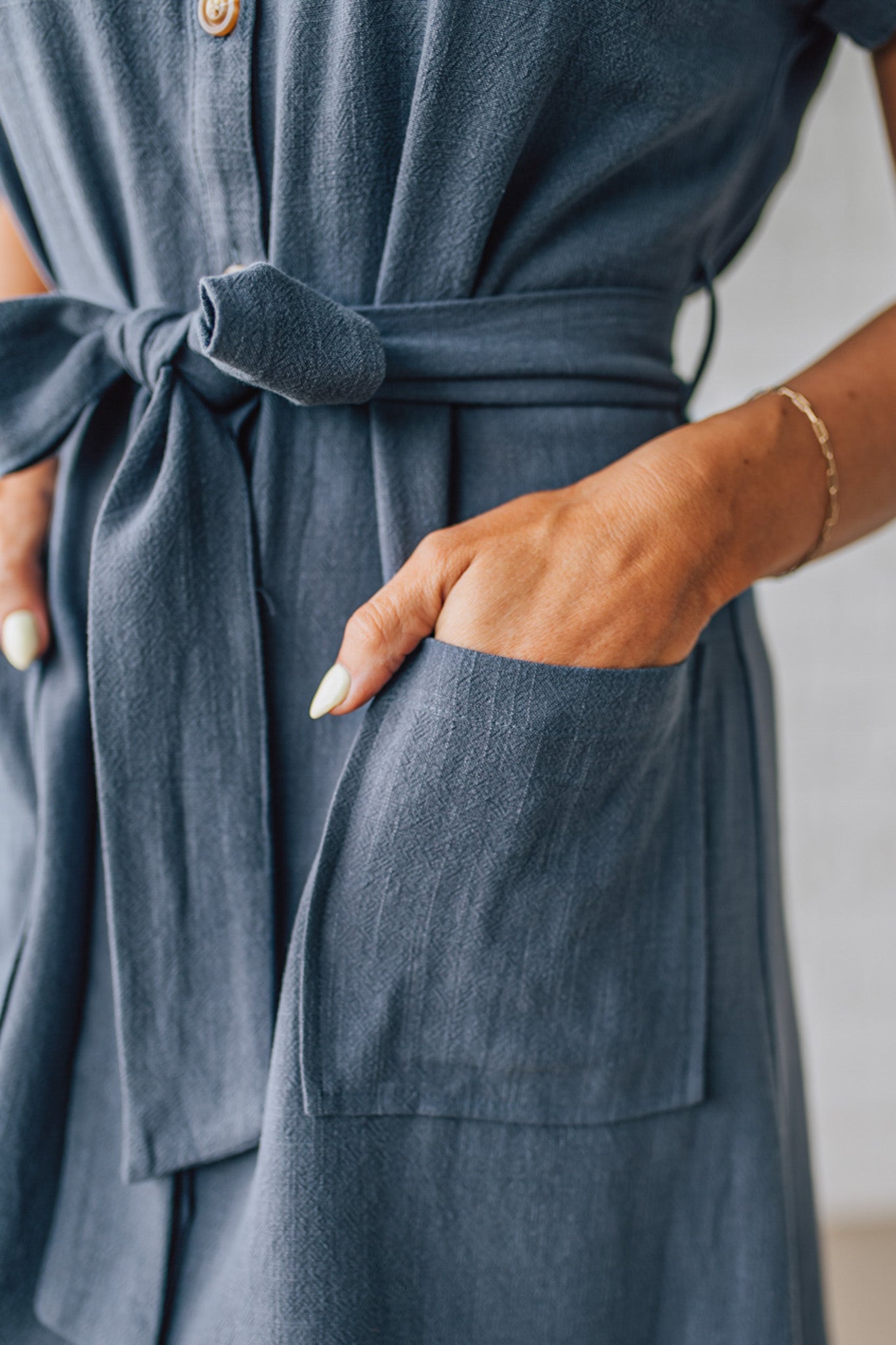 BLONDE WOMAN WARING A KNEE LENGTH SHORT DRESS WITH BUTTON UP AND COLLARED FRONT. HAS SHORT SLEEVES, FRONT PATCH POCKETS AND TIES AT THE WAIST, ALL IN SLATE BLUE WITH A TEXTURED LINEN BLEND.