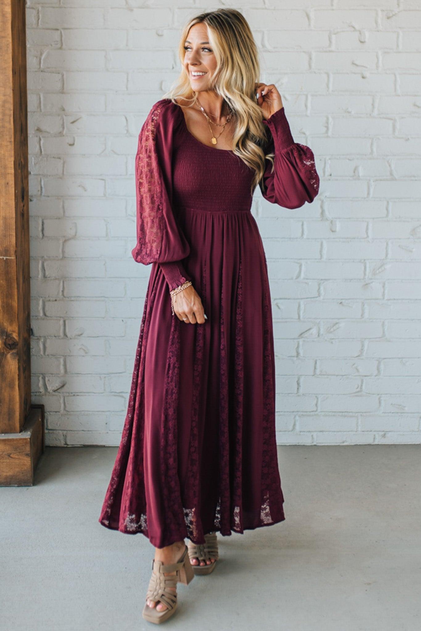 Woman wearing a burgundy dress with long sleeves against a white brick wall.
