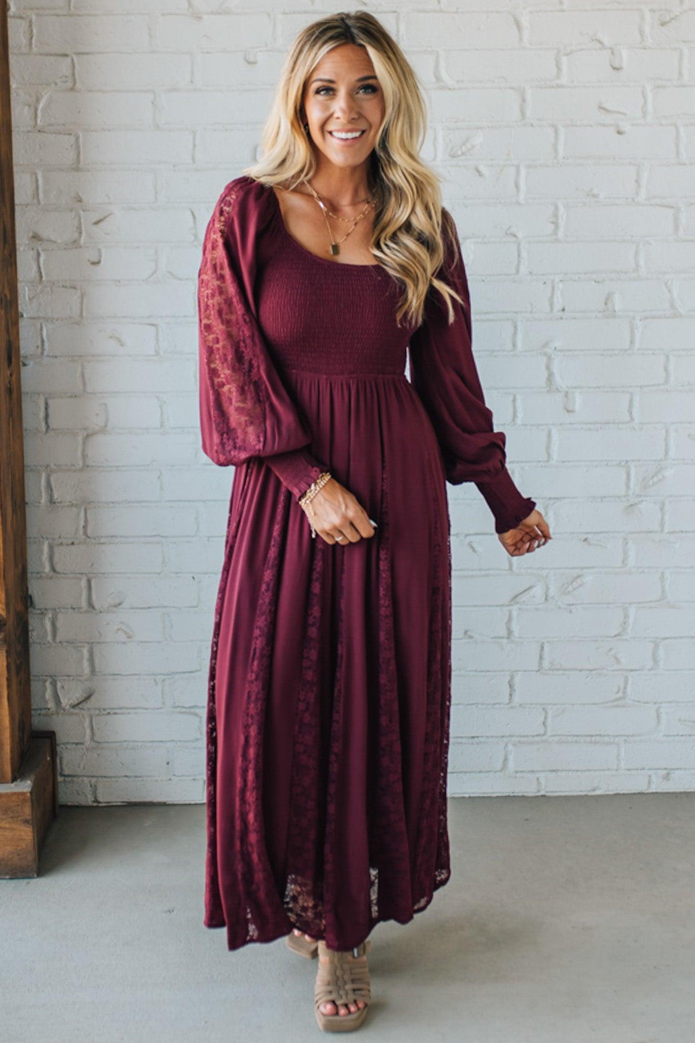 Woman wearing a burgundy dress with a matching cardigan against a white brick wall.