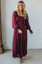 Woman wearing a burgundy dress with a matching cardigan against a white brick wall.
