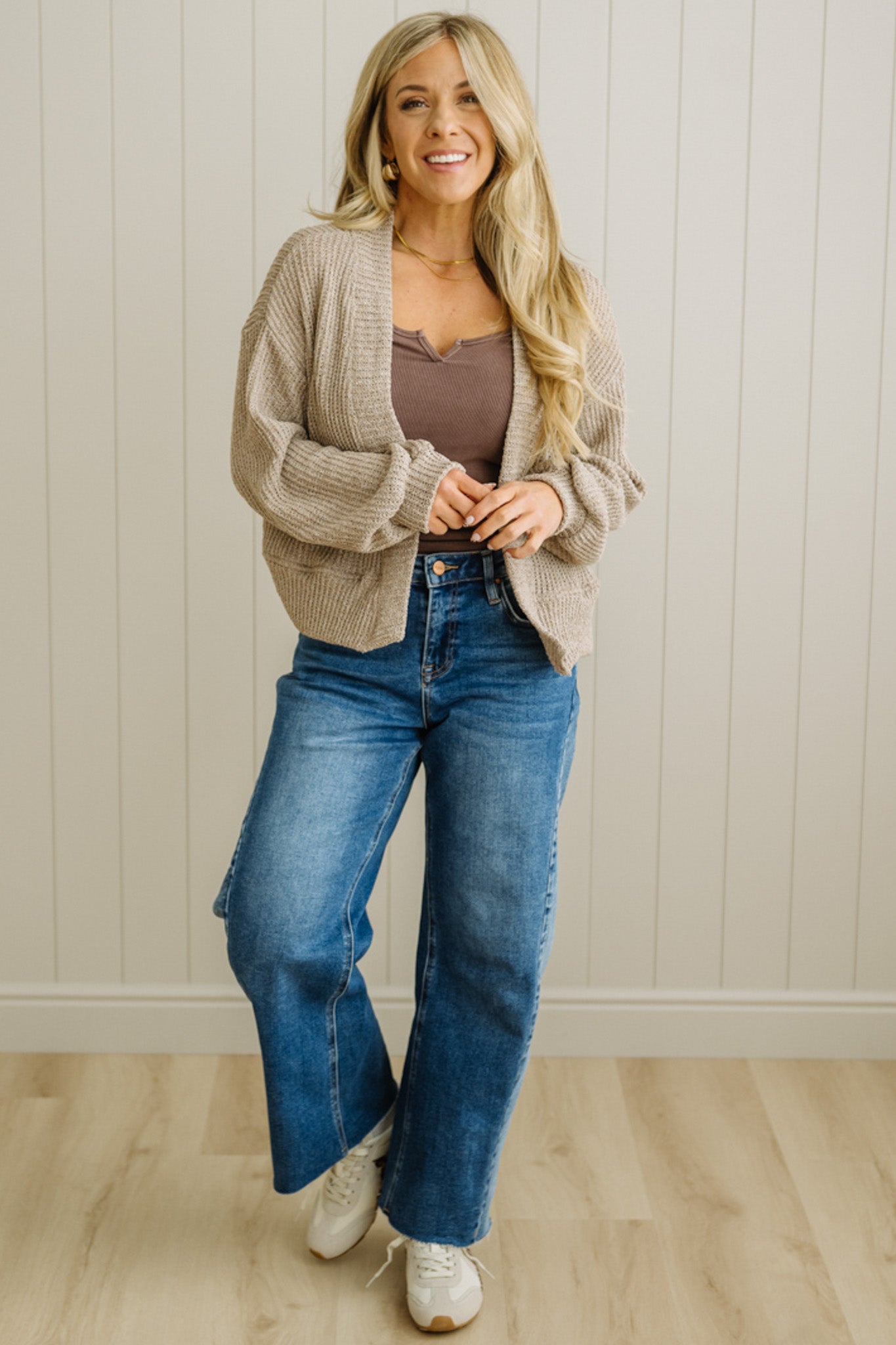 Woman wearing a beige cardigan, brown top, blue jeans, and white sneakers against a light wooden paneled wall.