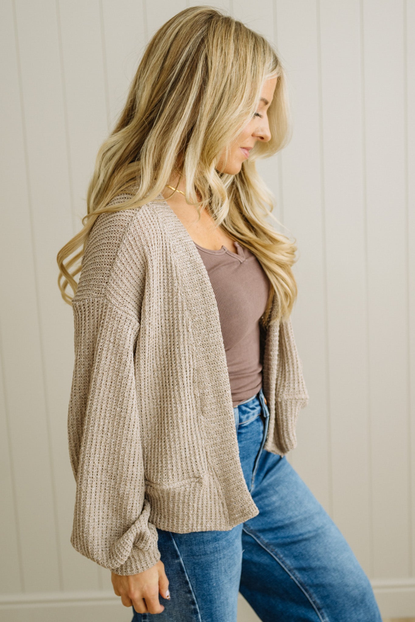 Woman wearing a beige cardigan, brown top, and blue jeans against a neutral background