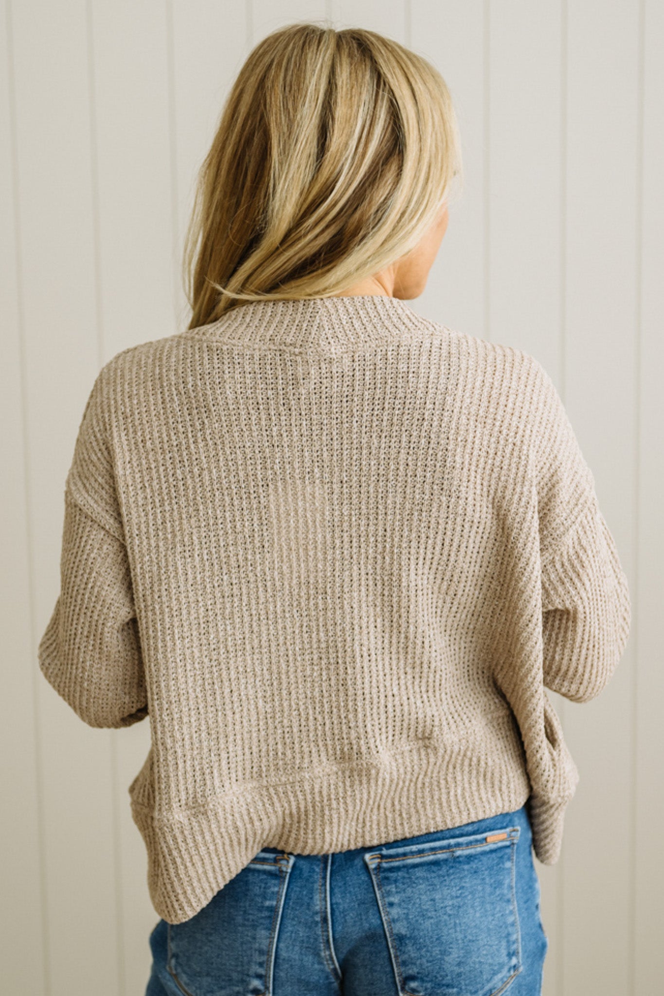 Person wearing a beige sweater and blue jeans against a light wooden panel background