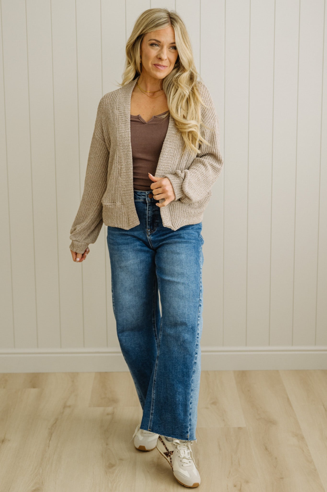 Woman wearing a beige cardigan, brown top, blue jeans, and white sneakers against a white paneled wall.