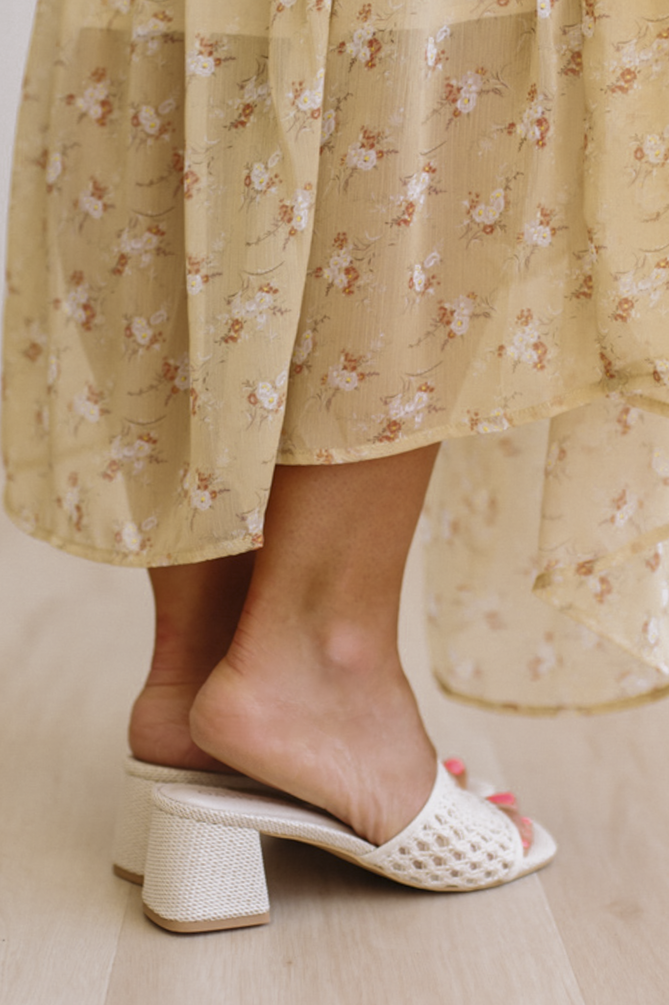 White open-toe sandals worn with a floral dress on a light wooden floor.