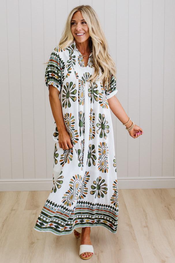 Woman wearing a long, patterned dress standing in a room with wooden flooring and a white wall.