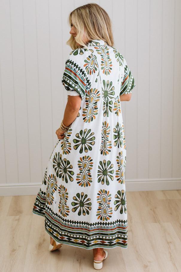 Woman wearing a patterned dress standing in a room with wooden flooring and white walls.