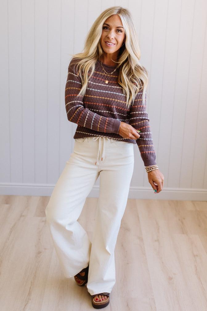 Blonde woman wearing an all over perforated mocha brown sweater with thin horizontal multi color stripes, a rolled neckline and ribbed cuff.