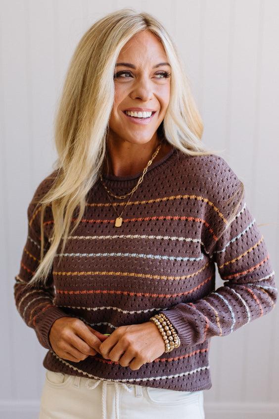 Blonde woman wearing an all over perforated mocha brown sweater with thin horizontal multi color stripes, a rolled neckline and ribbed cuff.