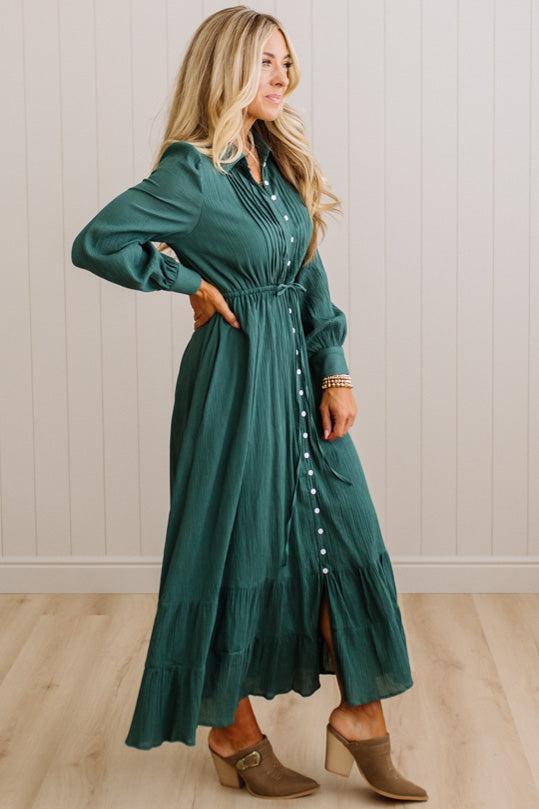 A woman modeling a long-sleeve, dark green midi dress with a button-down front, waist tie, and tiered skirt, paired with brown heeled mule boots against a white paneled wall and light wood floor.