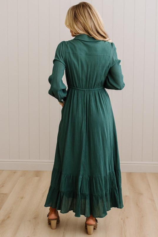A woman viewed from behind wearing a dark green long-sleeve midi dress with a gathered waist and tiered hem, standing on a light wood floor against a white paneled wall.