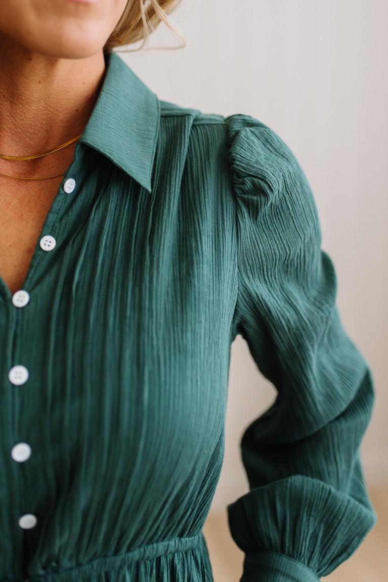 Close-up of a woman wearing a dark green crinkle-textured blouse with white buttons, featuring a pointed collar and puffed long sleeves.