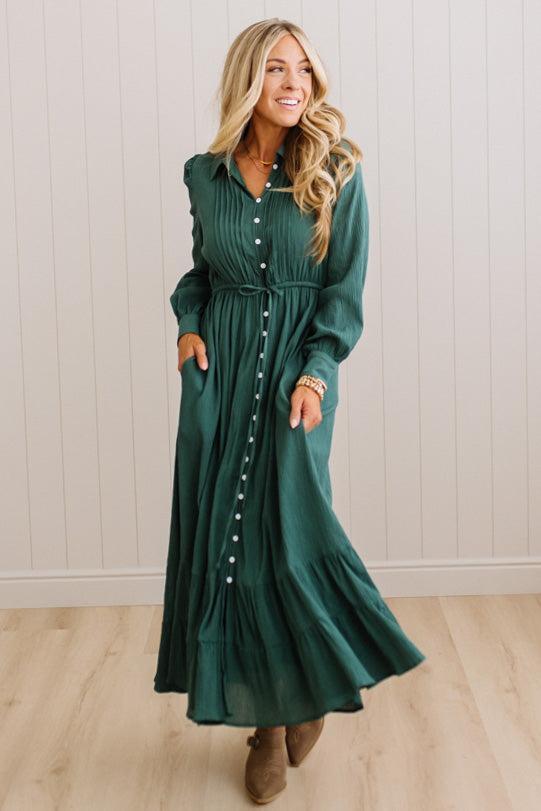 A woman wearing a long-sleeve, button-front maxi dress in dark green, standing in a room with light wood floors and white paneled walls.
