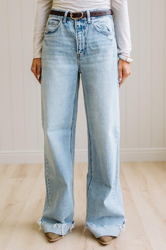 Woman wearing a gray t-shirt and light blue jeans standing on a wooden floor with a white wall background.