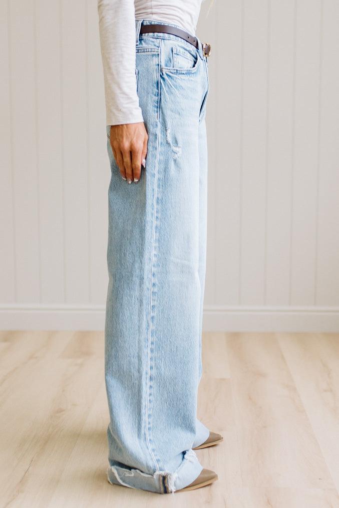 Woman wearing a gray t-shirt and light blue jeans standing on a wooden floor with a white wall background.