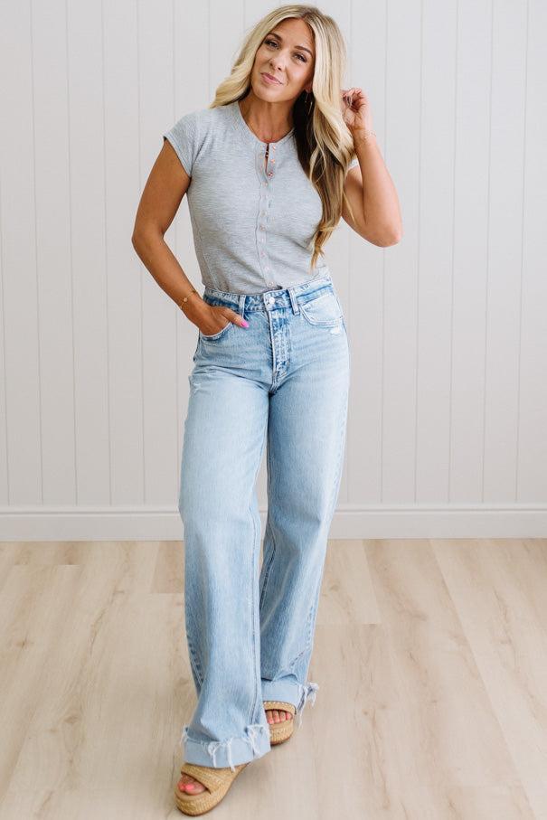 Woman wearing a gray top and light blue jeans standing against a white wall.