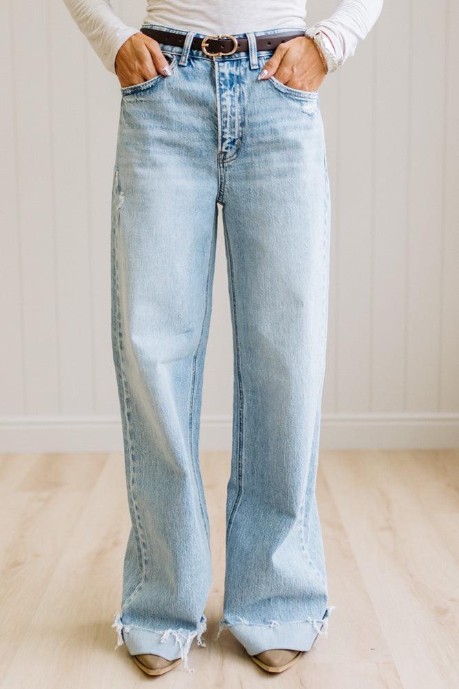 Woman wearing a gray t-shirt and light blue jeans standing on a wooden floor with a white wall background.