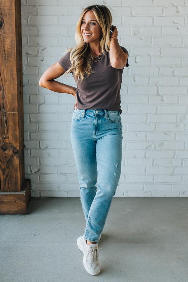 Woman wearing a brown t-shirt and light blue jeans standing against a white brick wall.