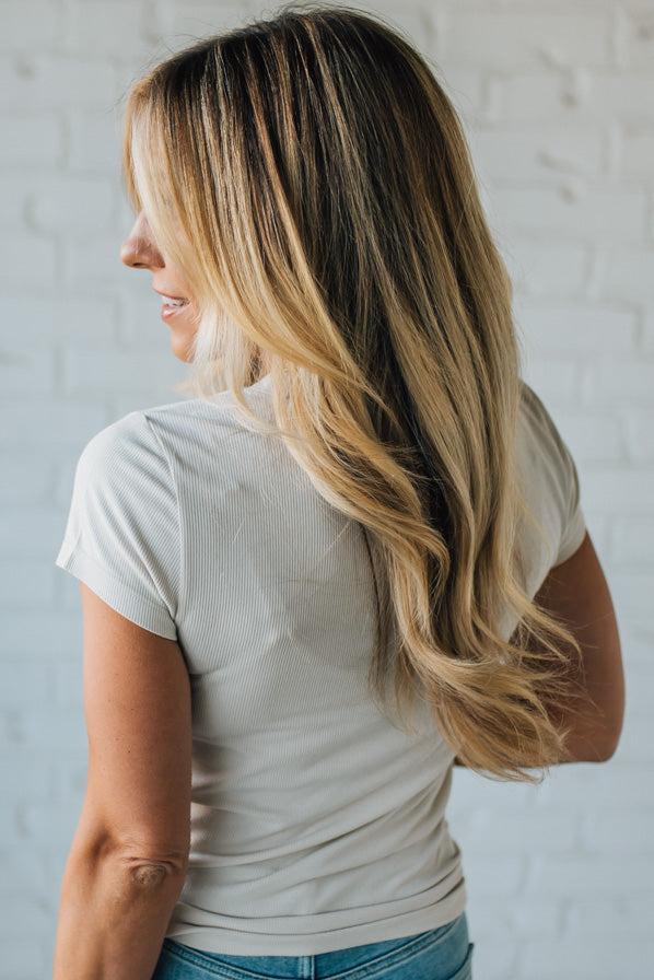 Woman with long, wavy hair wearing a light-colored top against a white brick wall.