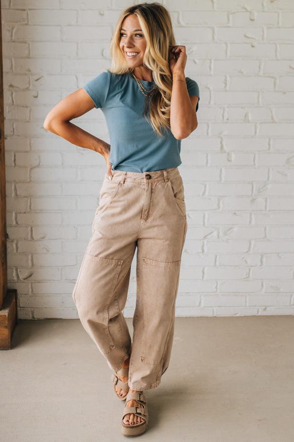 Woman wearing a blue shirt and beige pants standing against a white brick wall.