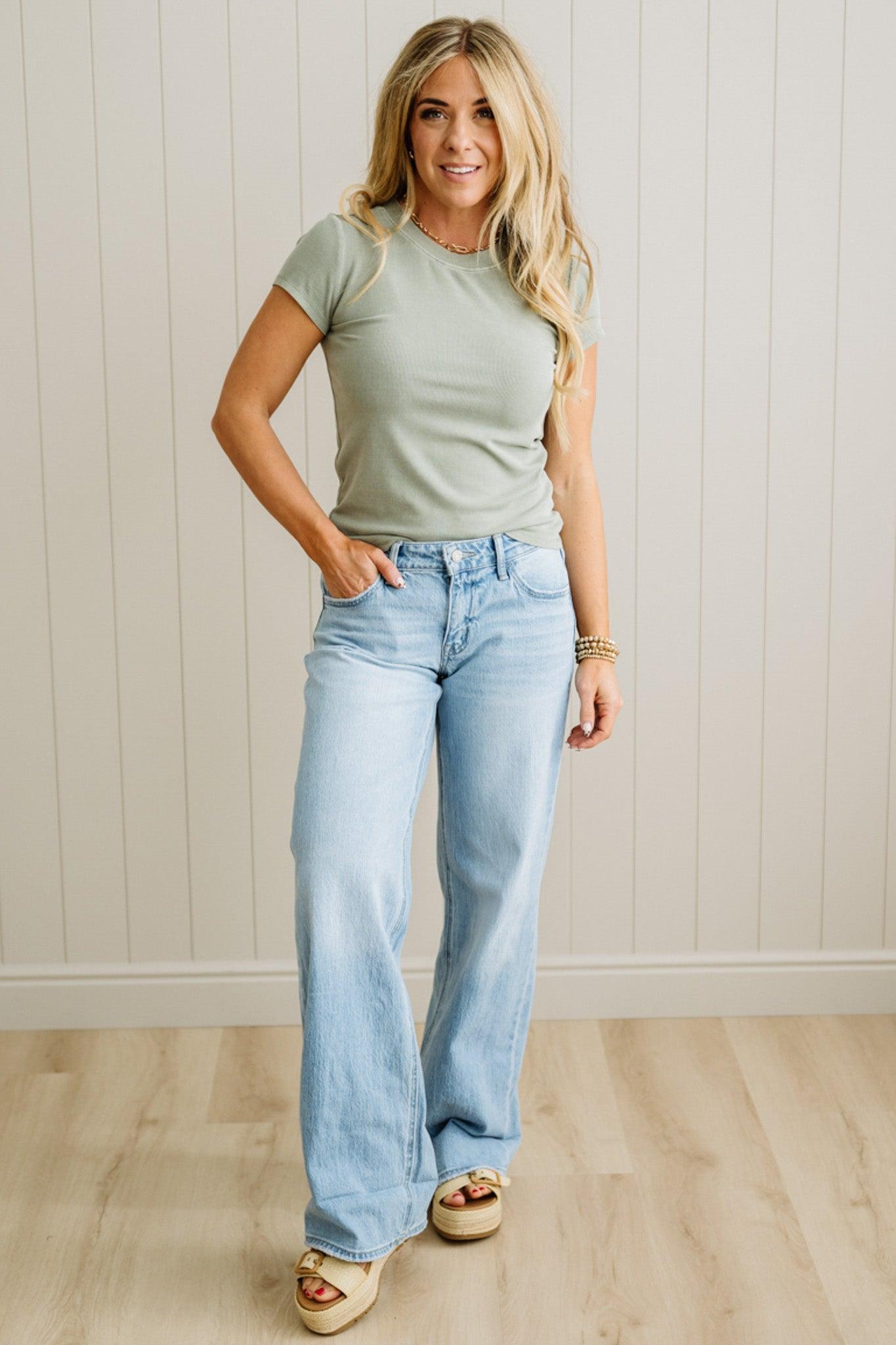 Woman wearing a green t-shirt and light blue jeans standing against a white paneled wall.