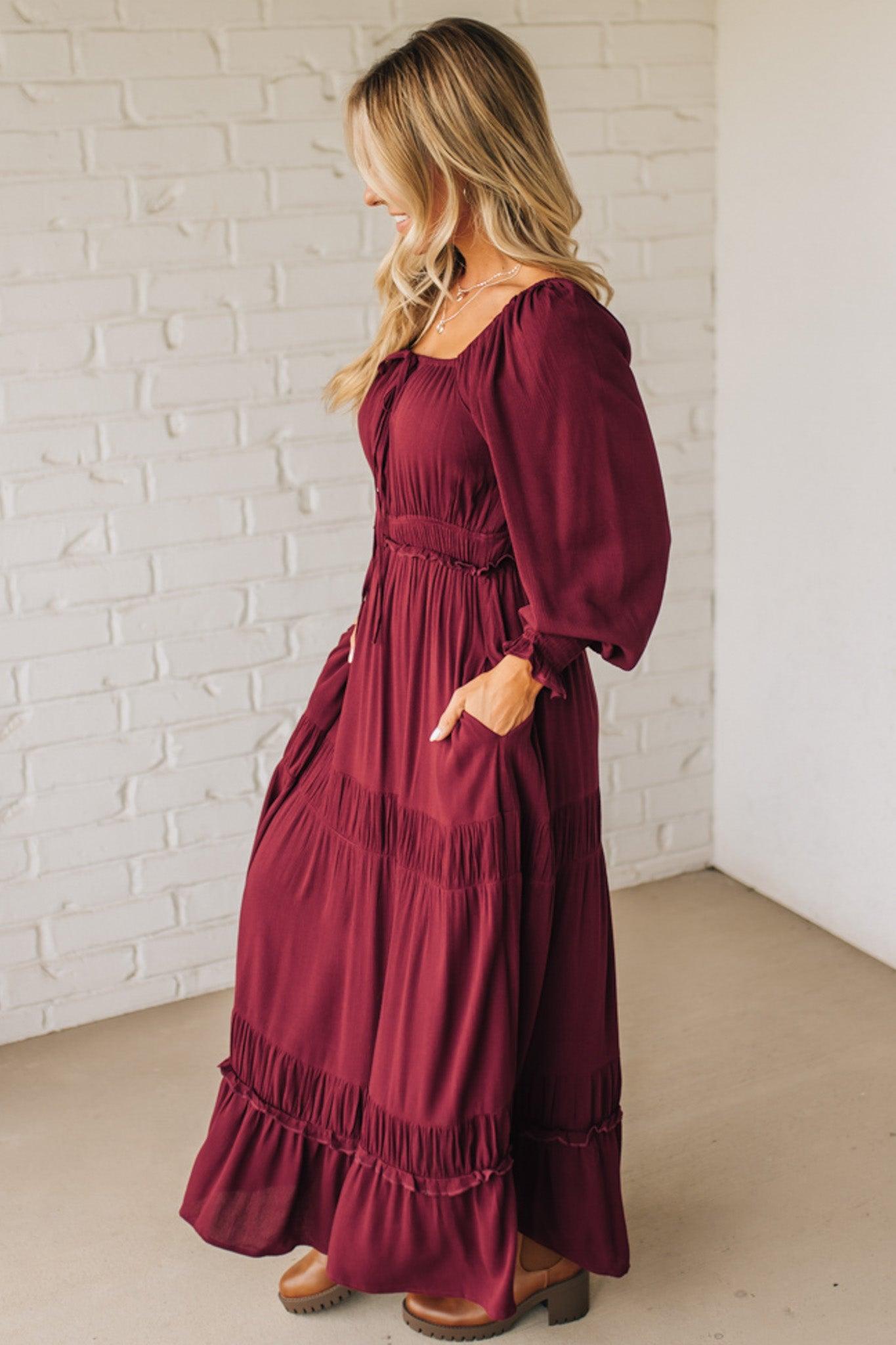 Woman wearing a burgundy dress with balloon sleeves against a white brick wall.