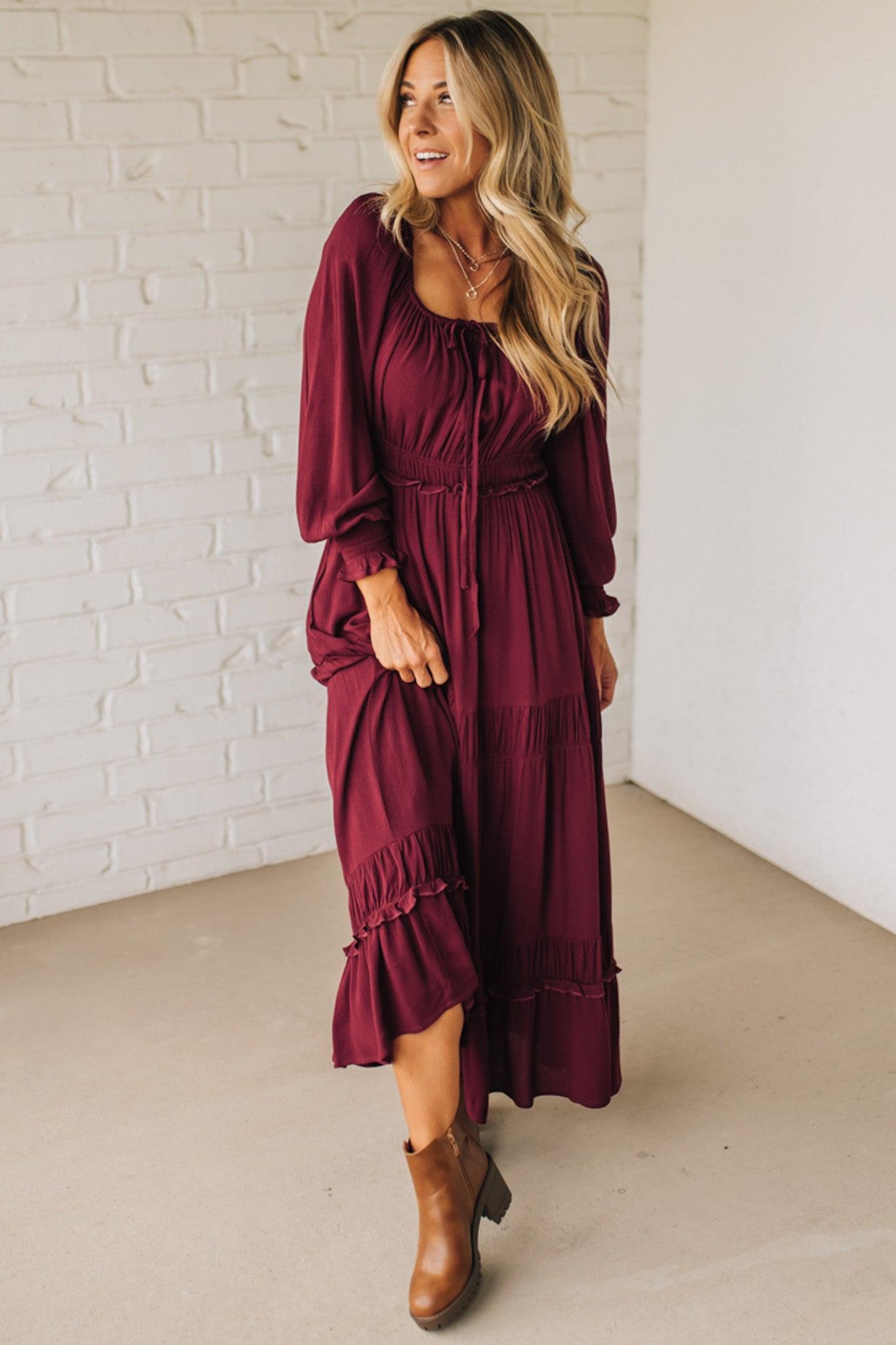 Woman wearing a burgundy dress with long sleeves and ruffled details, standing against a white brick wall.
