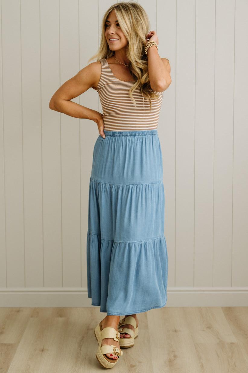 Woman wearing a beige top and blue tiered skirt against a light wooden paneled wall.