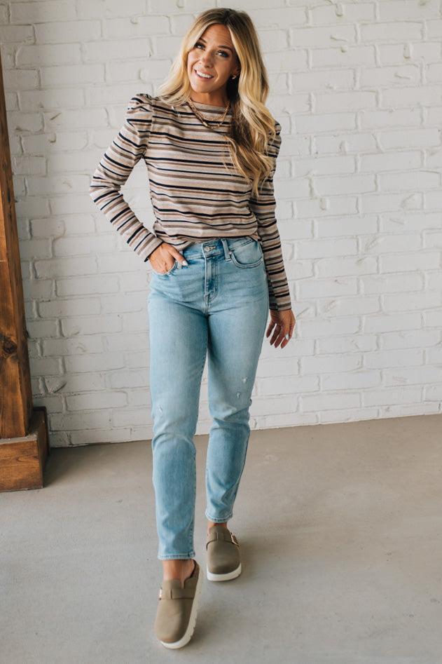 Woman wearing a striped long-sleeve top and light blue jeans against a white brick wall.