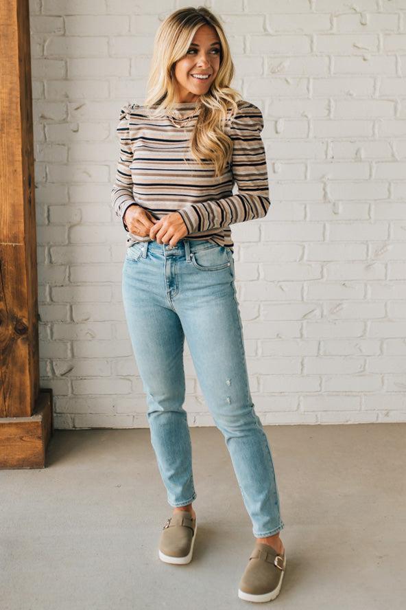 Woman wearing a striped sweater and light blue jeans standing against a white brick wall.