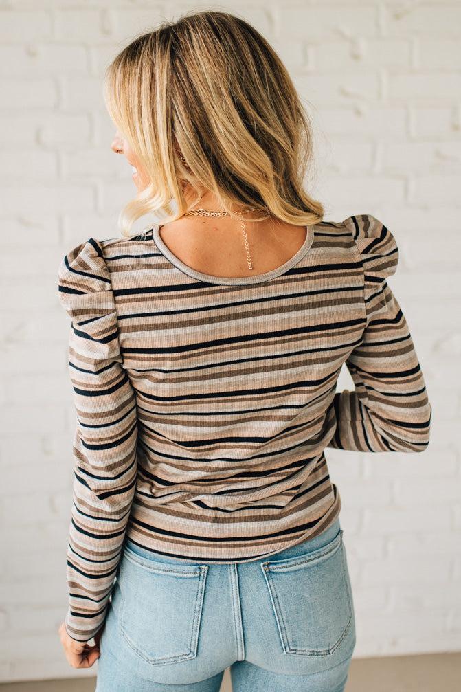 Woman wearing a striped long-sleeve top and light blue jeans against a white brick wall.