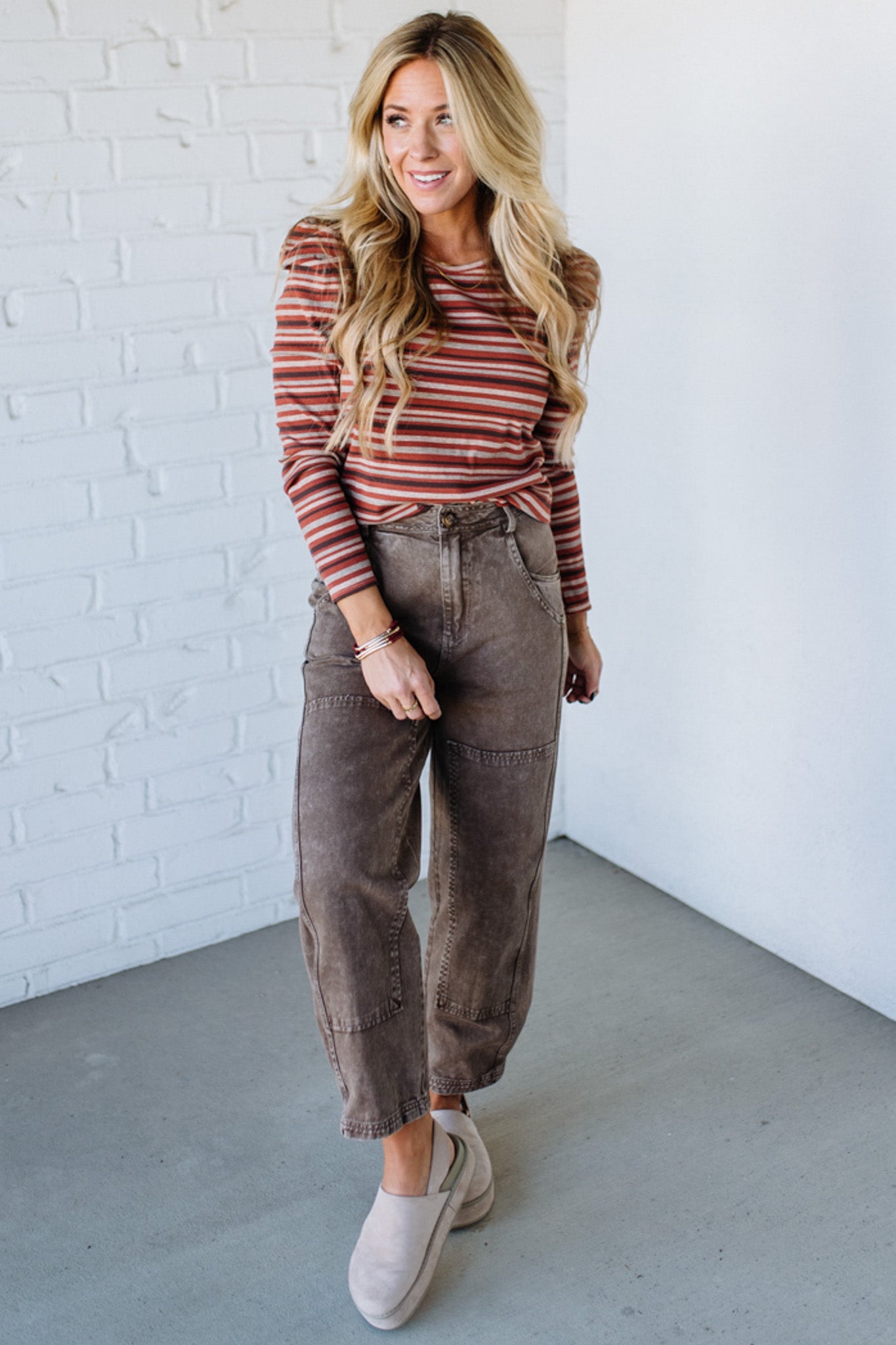 Woman wearing a striped shirt and brown pants against a white brick wall.