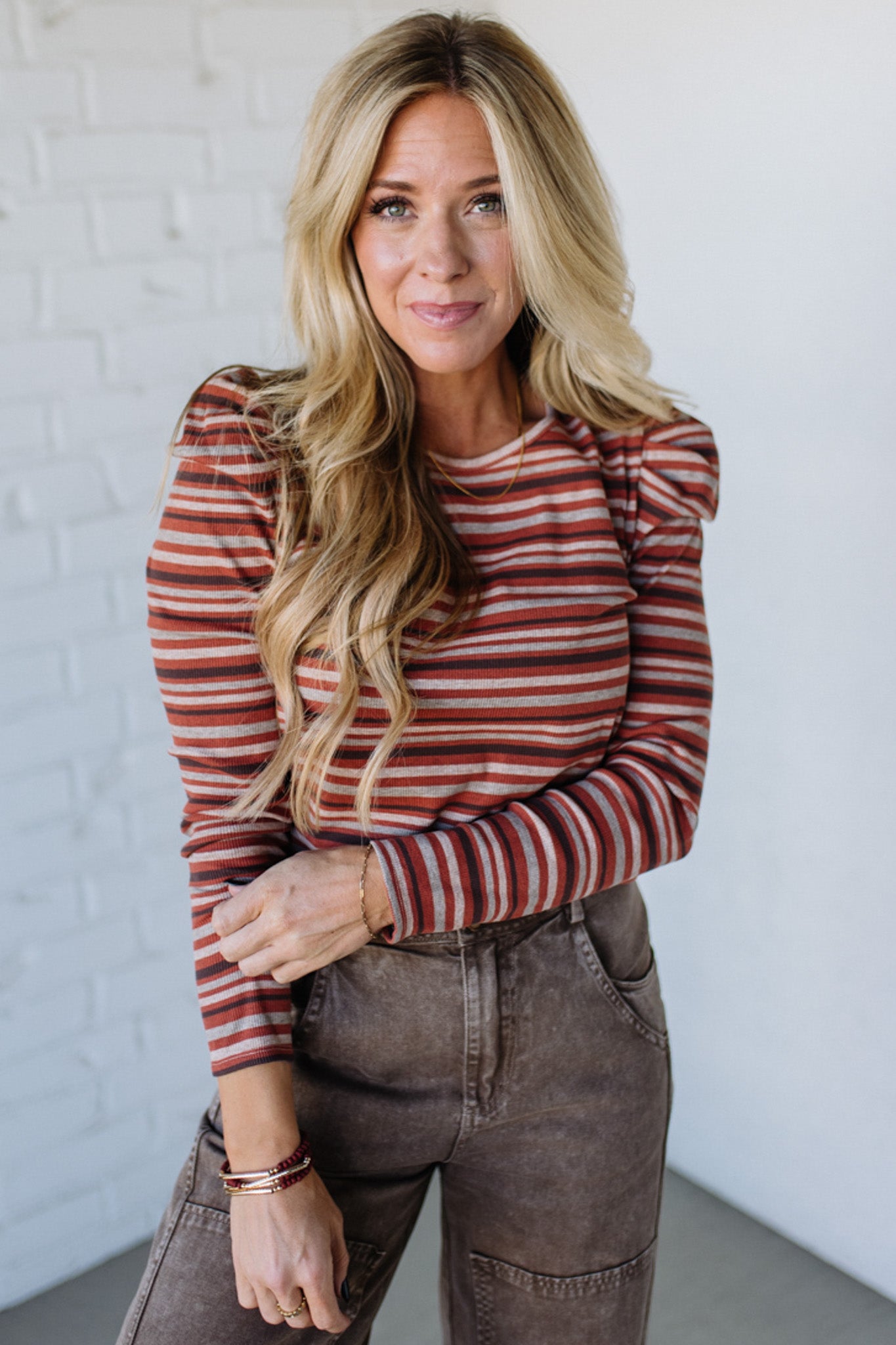 Woman wearing a striped sweater and brown pants against a white brick wall.