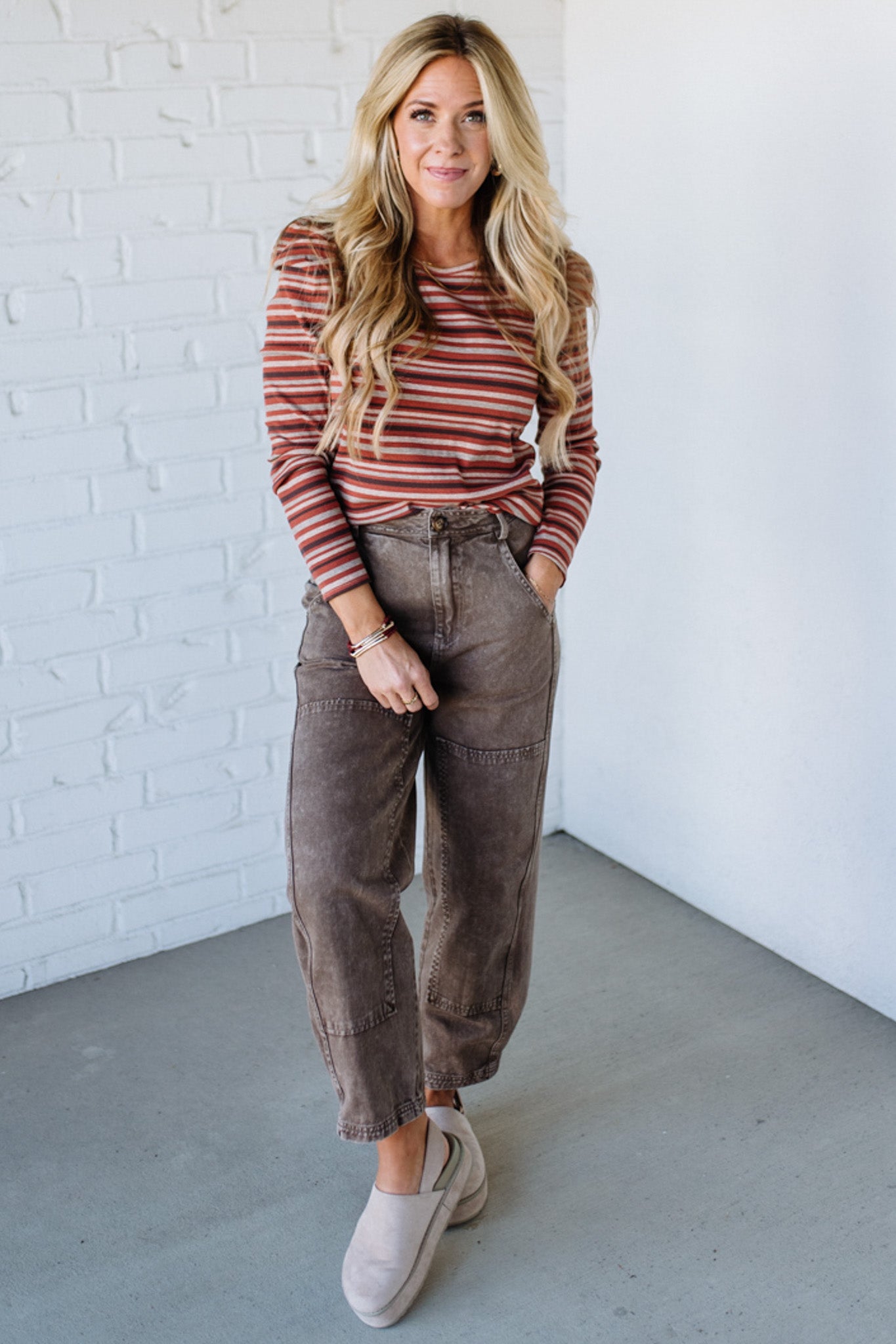 Woman wearing a striped sweater and brown pants against a white brick wall.