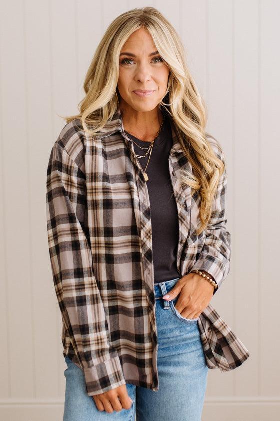 A woman modeling a long-sleeve plaid button-up shirt in brown, beige, and black, worn open over a black top and blue jeans against a white paneled wall.
