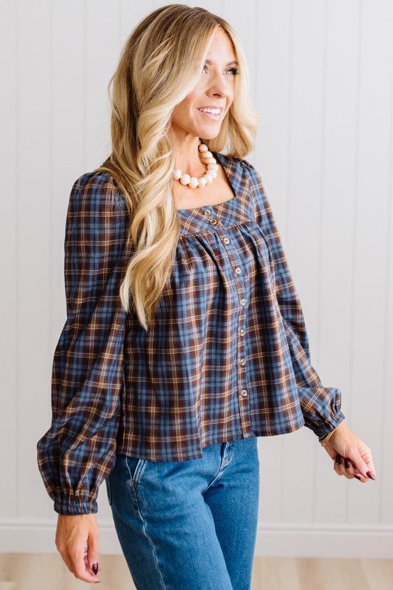Blonde woman in a brightly lit studio wearing a PLAID SQUARE NECK BUTTON CLOSURE FRONT LONG SLEEVE BLOUSE WITH DARK JEANS AND BROWN BOOTS.