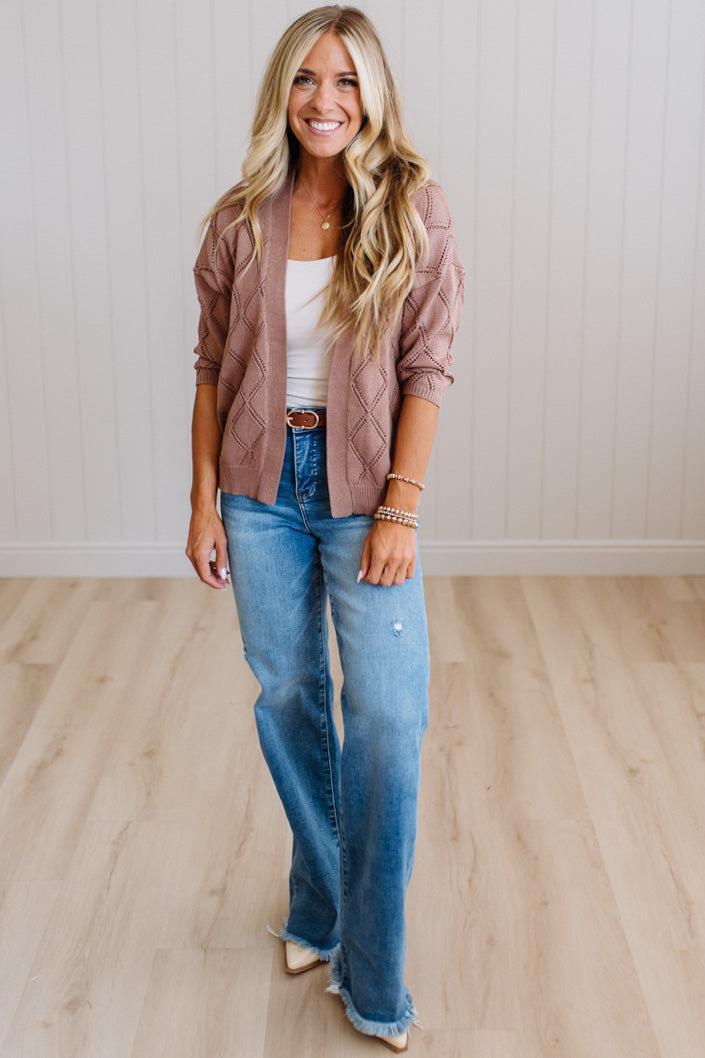 A woman stands in a bright room with light wood flooring and white paneled walls, wearing a dusty rose knit cardigan over a white tank top, blue jeans, and beige heels.