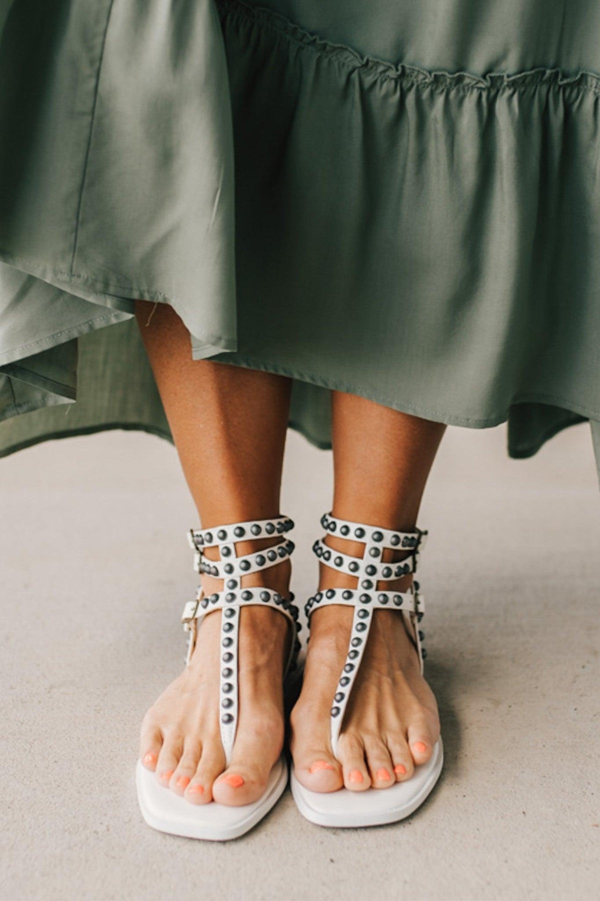 Woman in a green skirt wearing flat white faux leather gladiator sandals t-strap with triple buckle around ankle and pewter stud accents on straps.