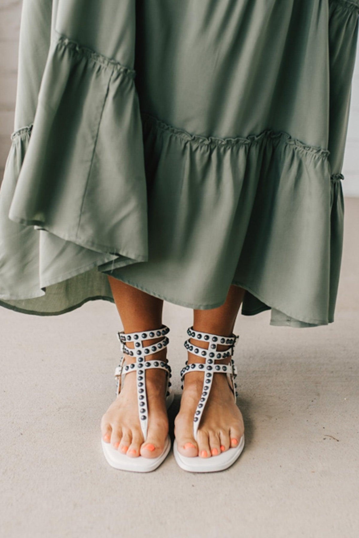 Woman in a green skirt wearing flat white faux leather gladiator sandals t-strap with triple buckle around ankle and pewter stud accents on straps.
