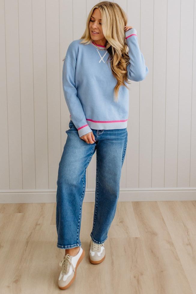 Woman wearing a light blue sweater with pink trim and jeans, standing against a neutral wall.