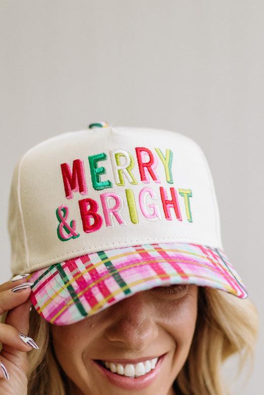 cream trucker cap pairs a crisp structured crown with a playful plaid brim in pink, green, and yellow, topped off with bold "Merry & Bright" embroidery in festive multicolor lettering