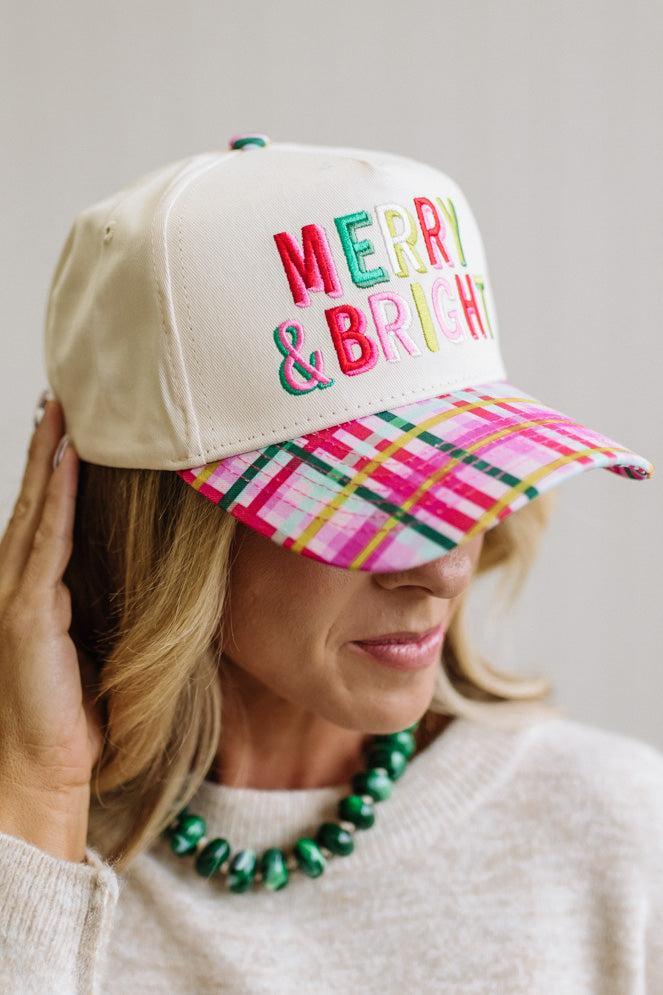 cream trucker cap pairs a crisp structured crown with a playful plaid brim in pink, green, and yellow, topped off with bold "Merry & Bright" embroidery in festive multicolor lettering