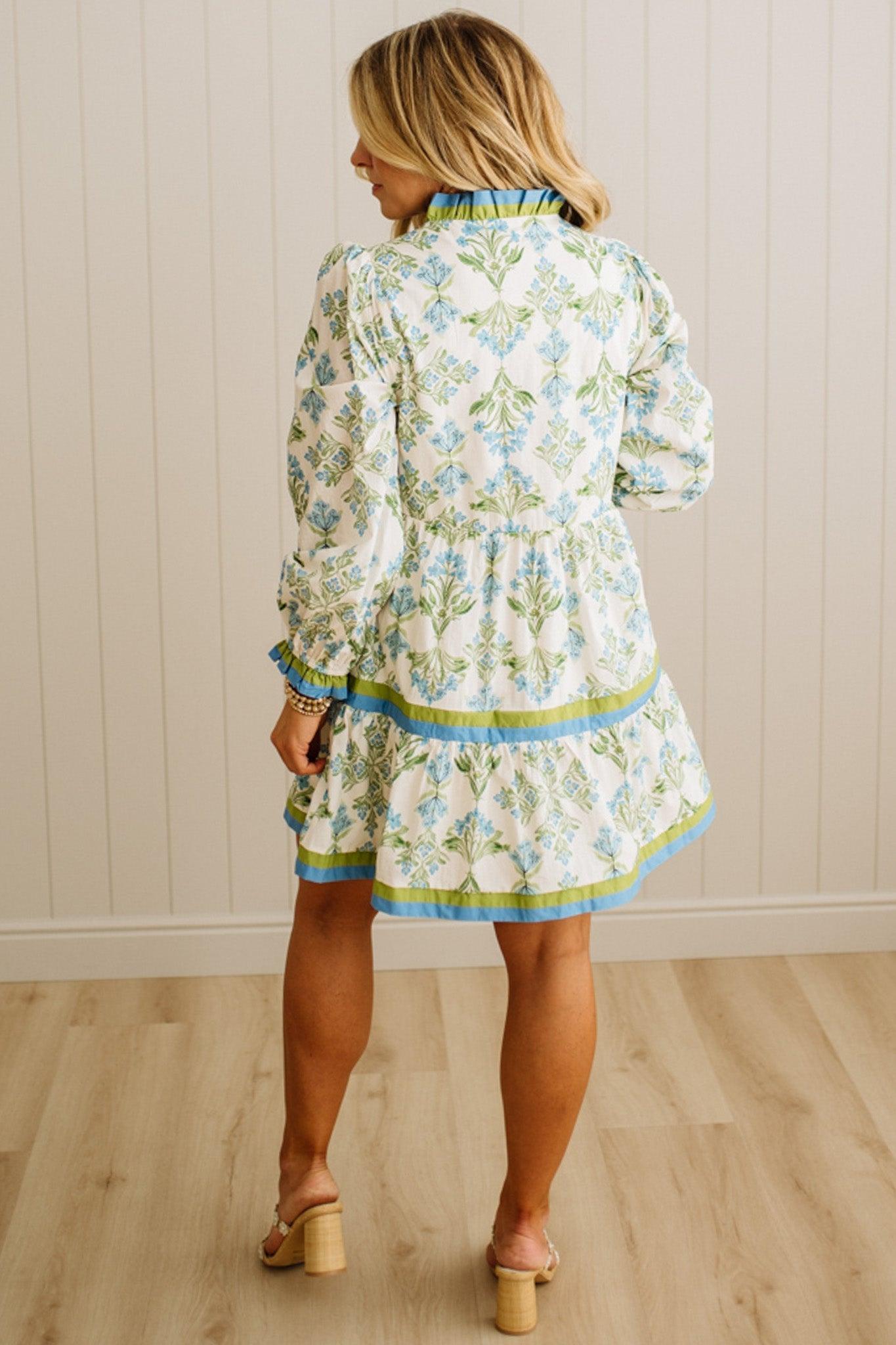 Woman wearing a floral dress standing against a light wooden paneled wall.